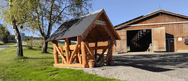 Rustikaler Holzpavillon mit Sitzgruppe, Blumenkübeln und Schild Heimatblick